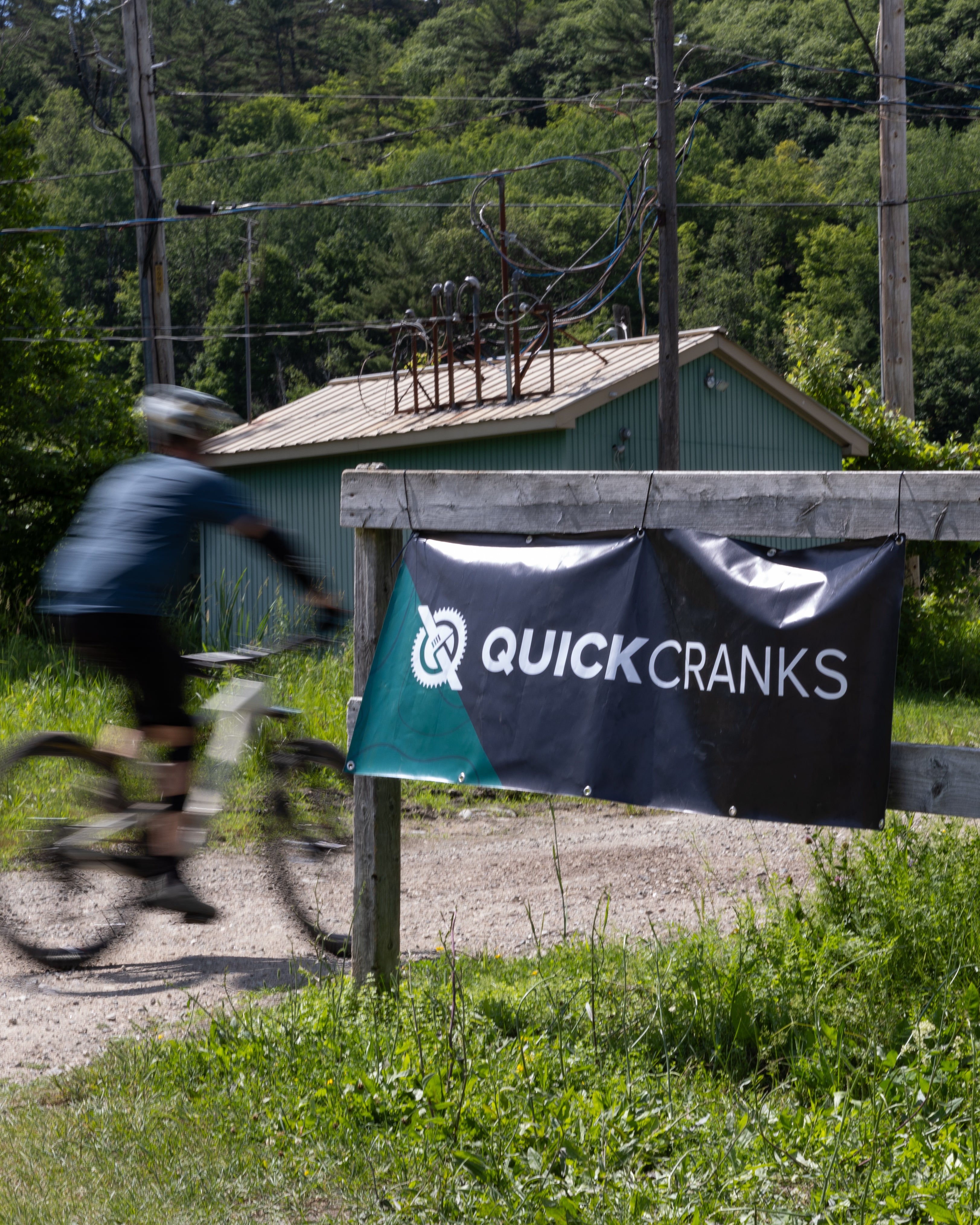 Person riding a bike near a building with a 'Quick Cranks' bike shop banner in a natural setting